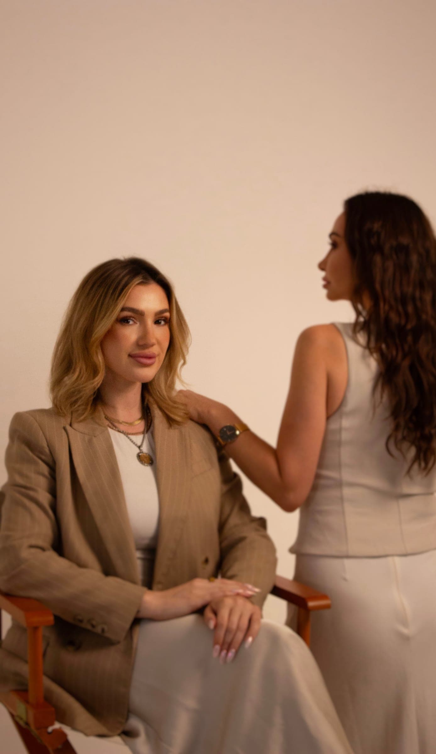 Ashley Sala seated in a tan blazer, with a teammate standing behind her on a blush backdrop