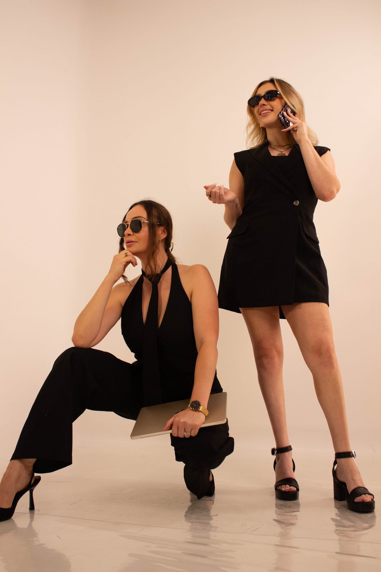 Ashley Sala and Megan Stagg in sunglasses on a blush backdrop, one crouching and one standing on the phone