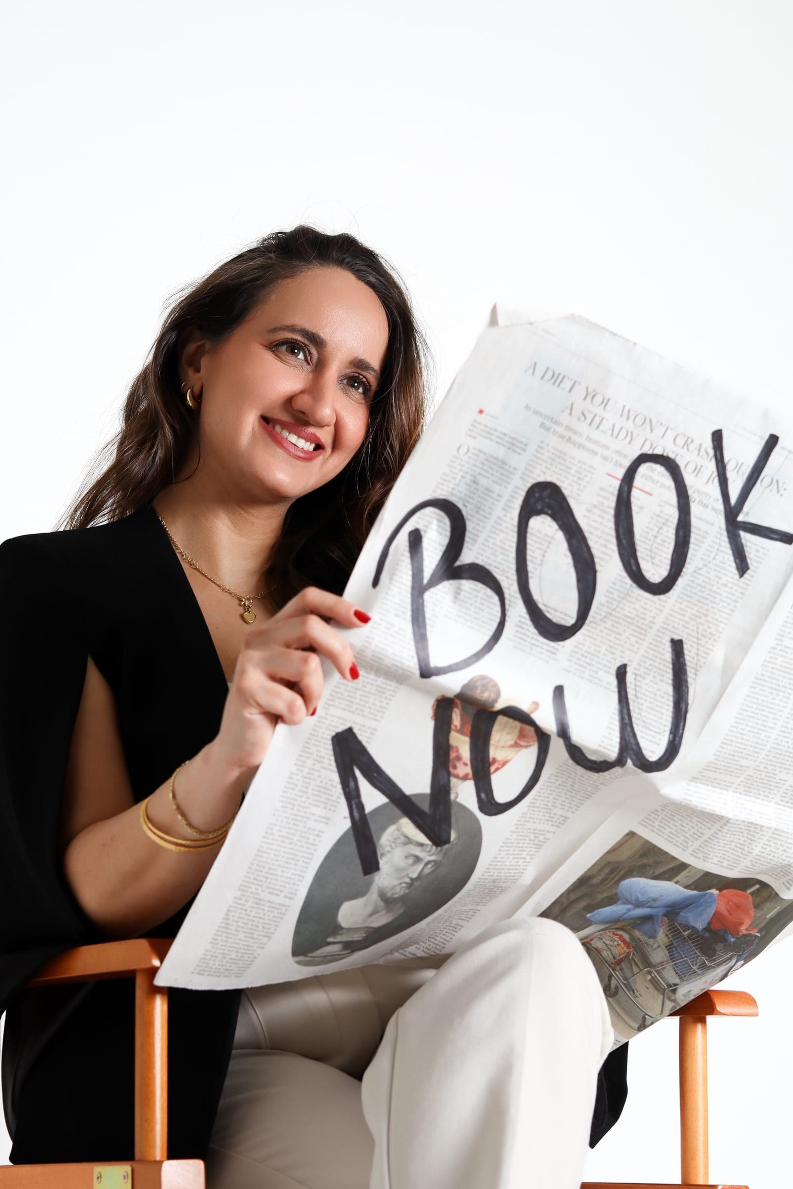 An agent seated in a director's chair holding a newspaper headed "Book Now"