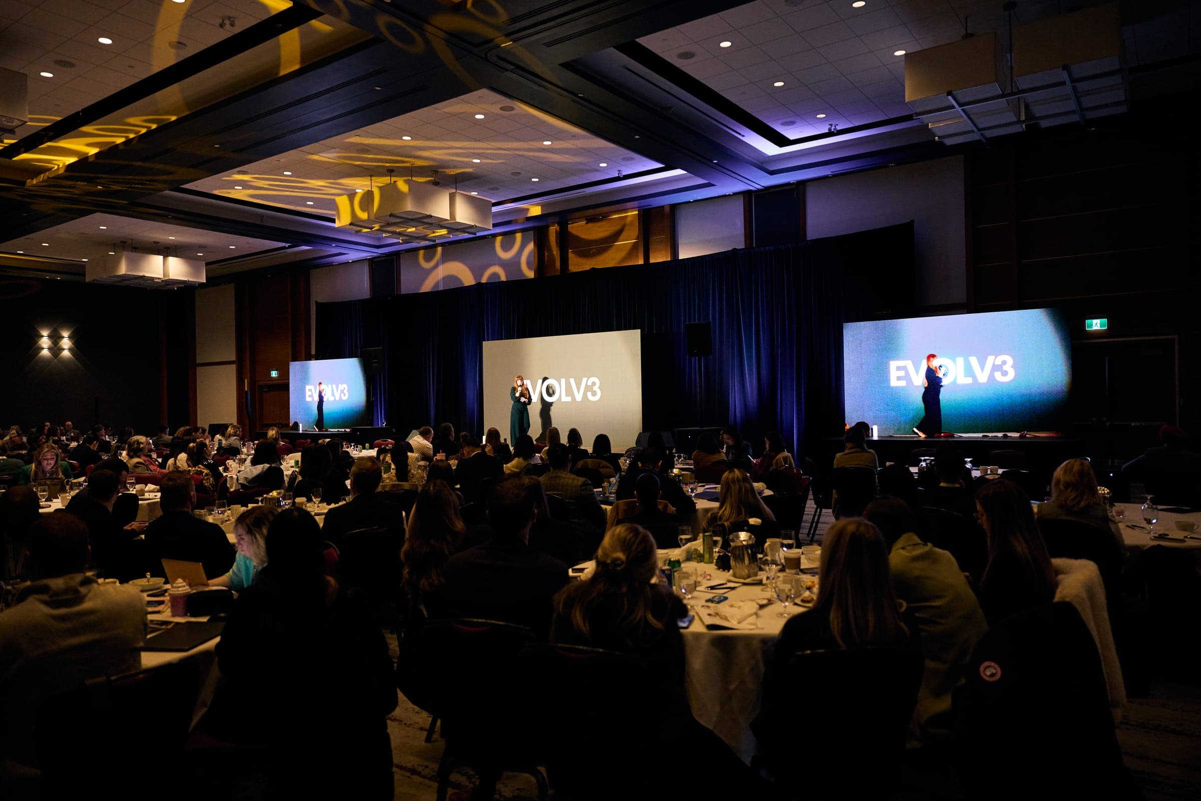 A packed ballroom during the Evolv3 conference, with the Evolv3 logo on the stage screens