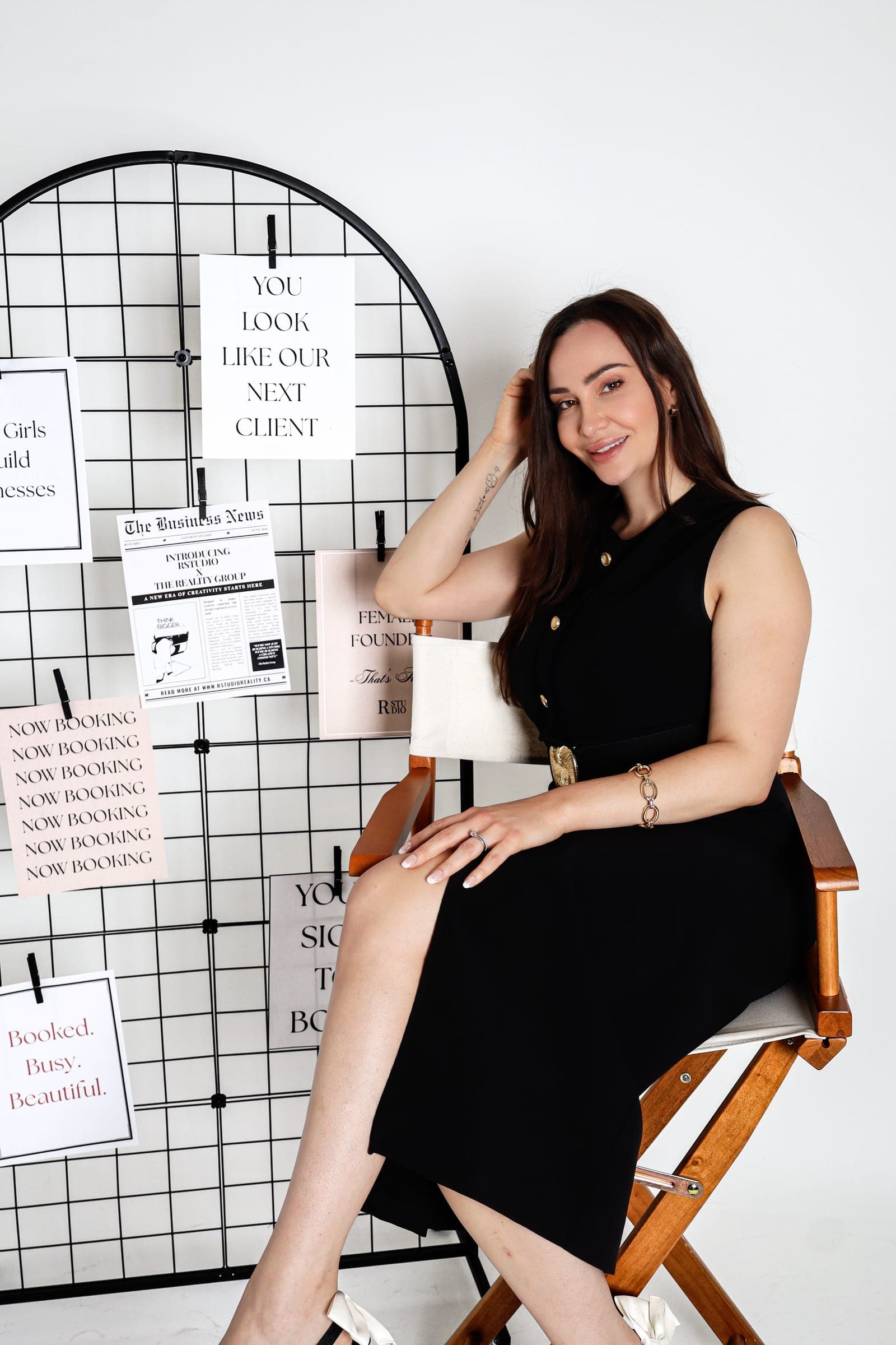 Megan Stagg, founder of The Reality Group, seated in a director's chair in a black dress beside a wire grid hung with prints