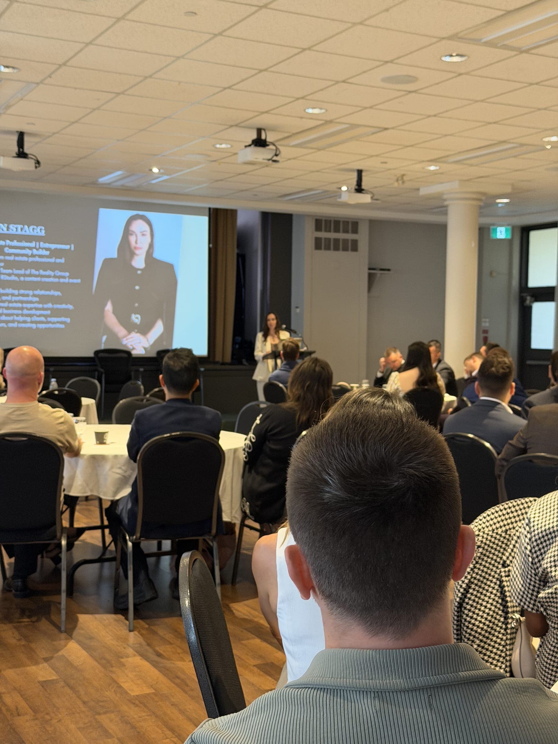 A speaker presenting to a seated audience in a meeting room, with her image on the projector screen