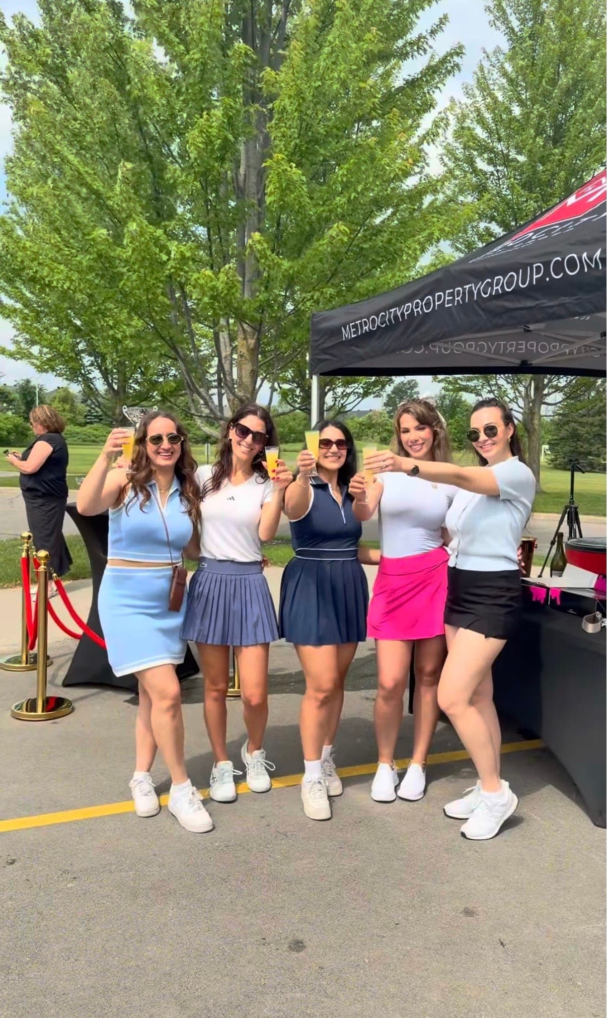 Five women in matching summer golf outfits raising drinks in front of a Reality Group tent at a golf day
