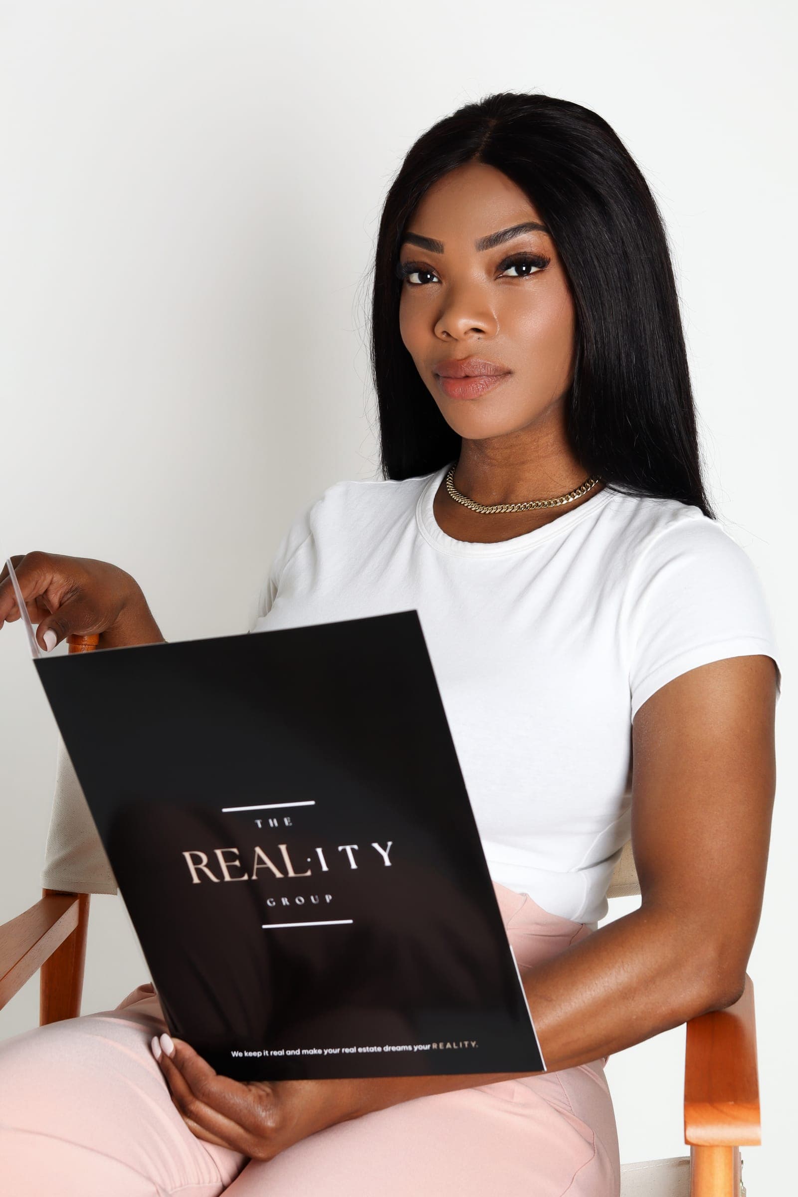 Noellee seated in a director's chair in a white tee, holding a black Reality Group folder