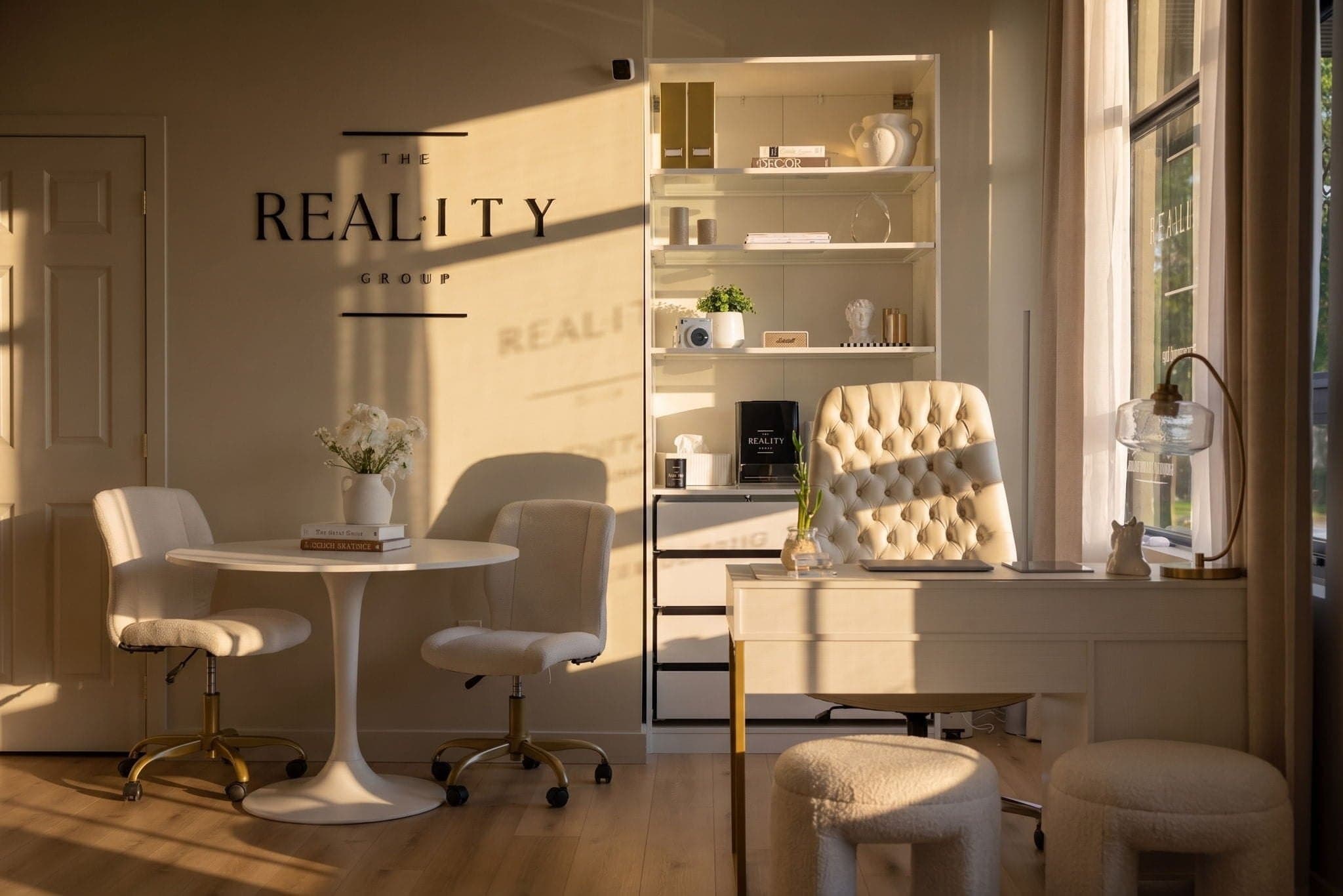 The Reality Group sign on a sunlit office wall, beside a round table with two chairs, a tufted desk chair and styled shelves