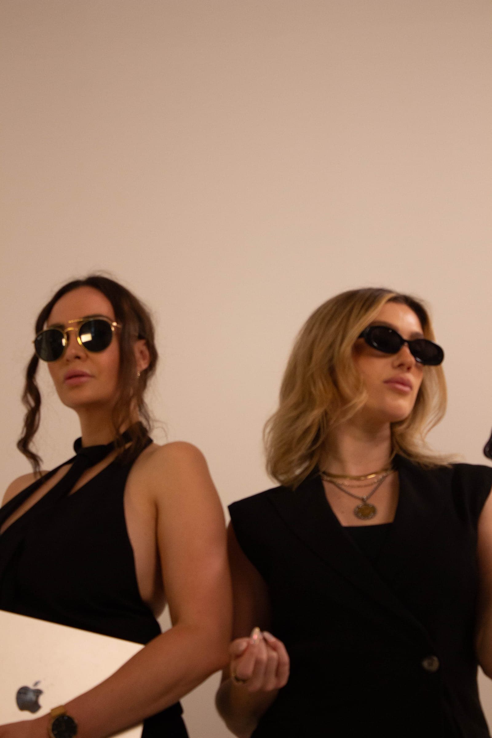 Two agents in sunglasses on a blush backdrop, one holding a laptop
