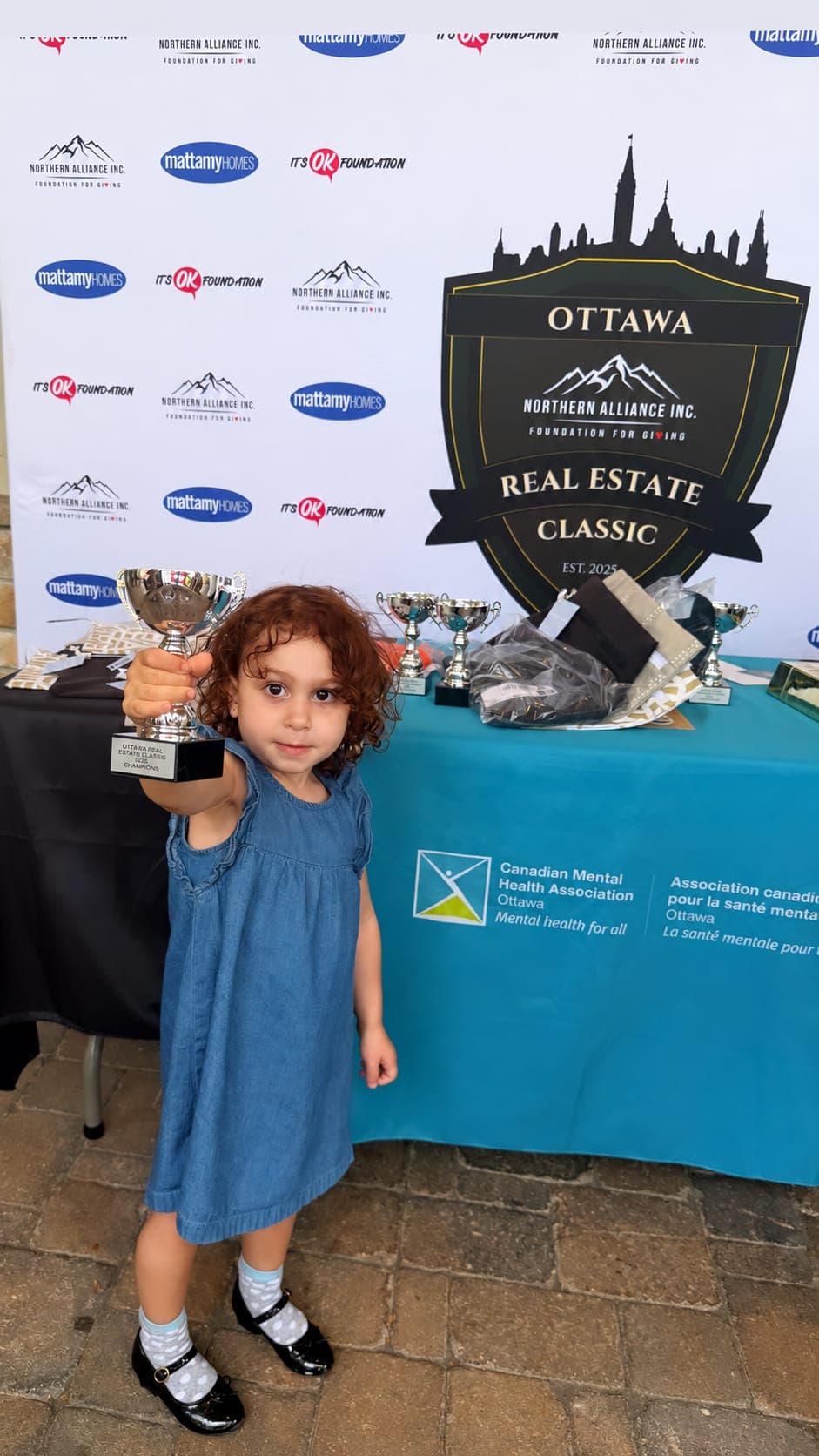 A small child in a denim dress holding up a silver trophy in front of an Ottawa Northern Alliance Real Estate Classic backdrop